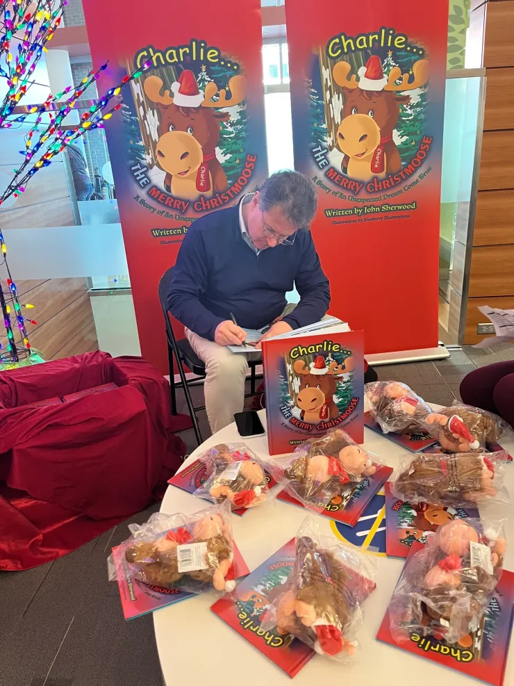 Man sitting in front of two “Charlie the Merry Christmas Moose” banners, signing a book at a table with plush moose toys and copies of the book.